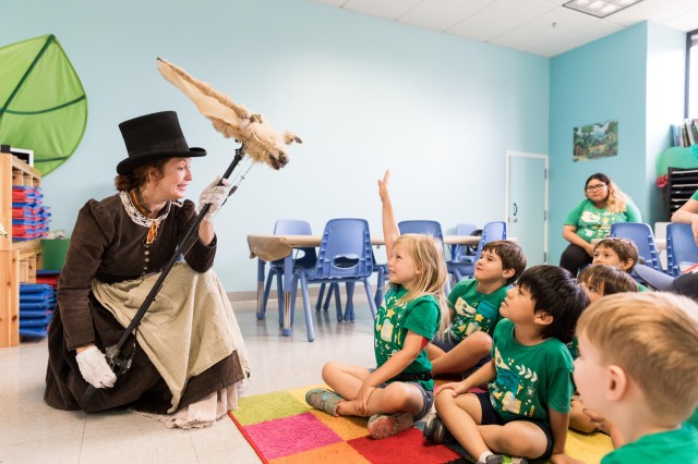 campers sit on the floor of a classroom, one girl raises her hand, while an adult uses a marionette puppet of a pterosaur