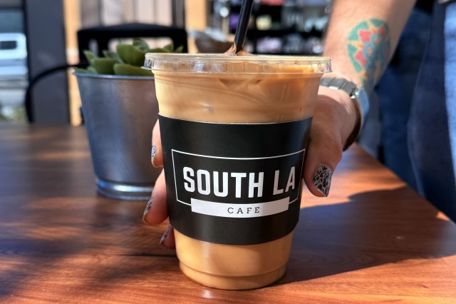 Iced coffee in a clear plastic cup with a black straw and a black label with the logo "South LA Cafe". A woman's hand reaches for the cup on a wooden table.