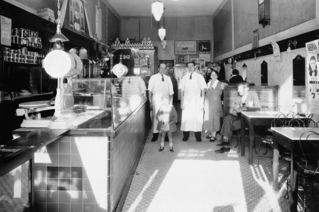Black and white, 1930s photograph of a multi-generational family in chef aprons standing inside their family deli.