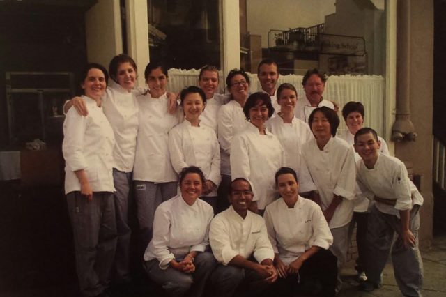 Sixteen adult men and, women wearing chef coats, posing for a class photo at a culinary arts school.
