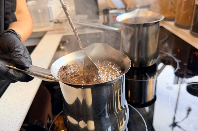 Close-up of a person wearing a black glove stirring a bubbling mixture in a small metal saucepan on a stovetop, with steam rising and another pot heating in the background.