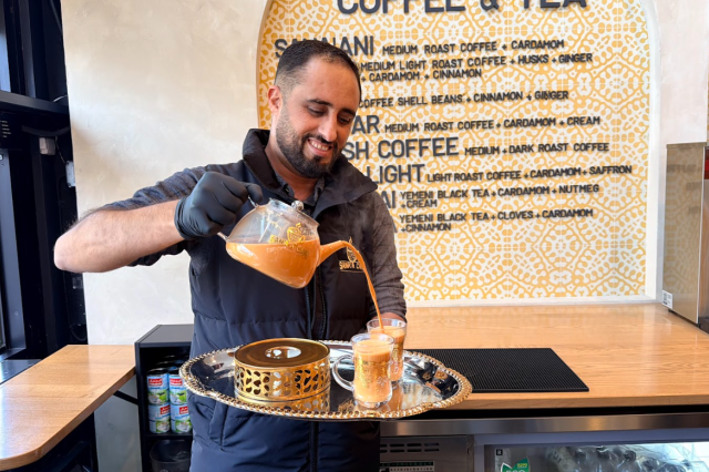 A barista wearing a glove pours a steaming, creamy drink from a glass teapot into small glass cups on a tray, smiling behind a café counter with a menu displayed on the wall.