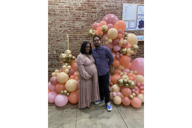 A woman and man stand in front of a colorful gold, orange and pink balloon arch. 