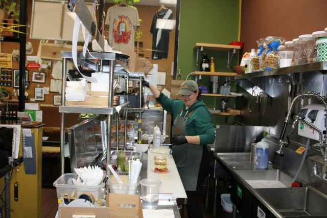 Michaela, a woman in a green sweater, cap and a gray apron, makes food orders in the kitchen section of the cafe.
