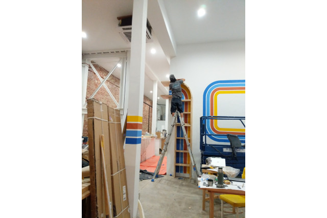 A man is putting up a corner shelving unit while standing atop a ladder in a coffee shop still being set up. 