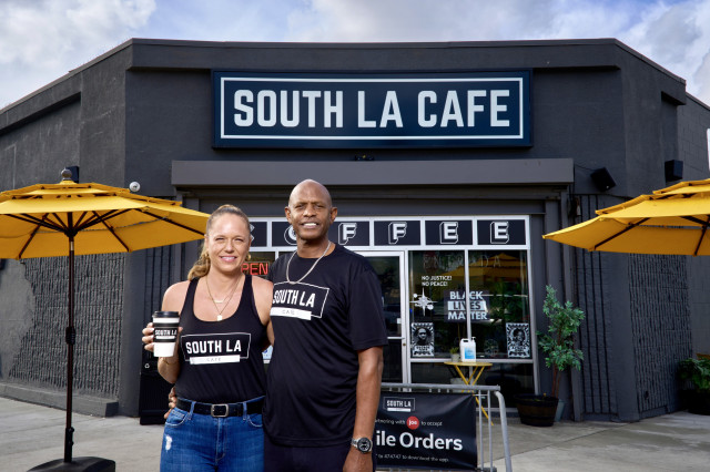 Man and woman standing in front of a grey-colored corner coffee shop with the sign "South LA Cafe" above the door. They are both wearing t-shirts with the cafe logo on them; the woman is holding a coffee cup.