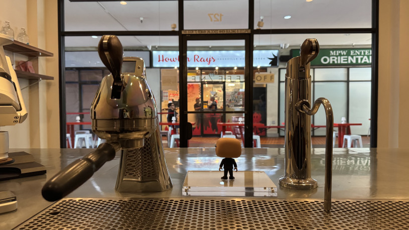 Interior of a coffee shop looking over the bar, past the espresso equipment, out to the glass entryway beyond.