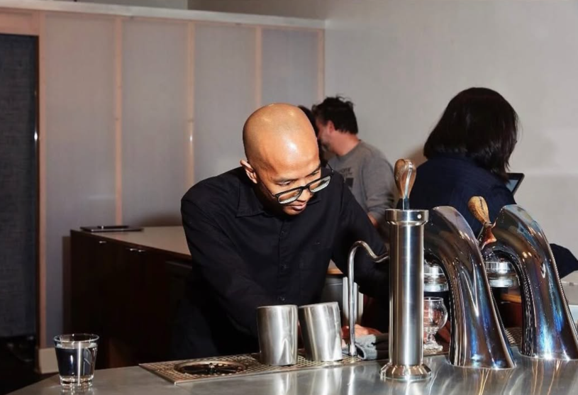Two baristas behind a coffee bar making drinks in front of customers in the background.
