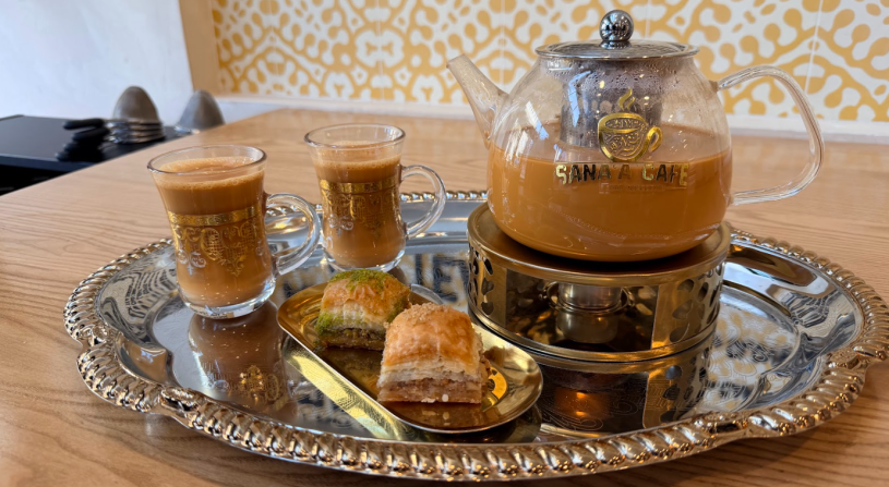A glass teapot filled with milky chai tea sits on a small warmer, accompanied by two clear glass cups of tea on a metal tray. In the background, a menu lists options like Aden chai and black tea with spices such as cardamom and cloves.