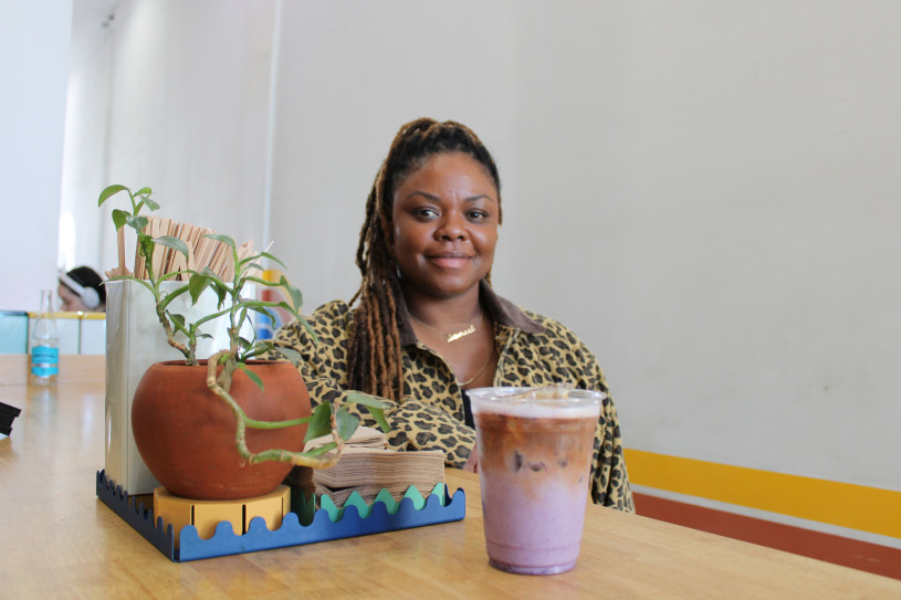 A woman in long braids and a leopard print jacket poses at a coffee counter with a purple ube latte in front of her.