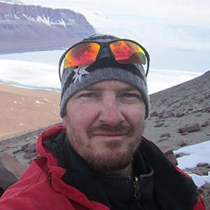 Nate Smith wearing a hat and goggles on his head with mountains, ocean, and beach in the background