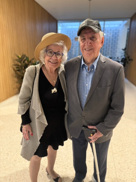 An elderly Armenian couple stand side by side, hugging. The woman dons glasses, a straw sunhat, a black dress, and a beige cardigan. The man sports a gray suit, with a blue shirt, and a gray baseball cap. 